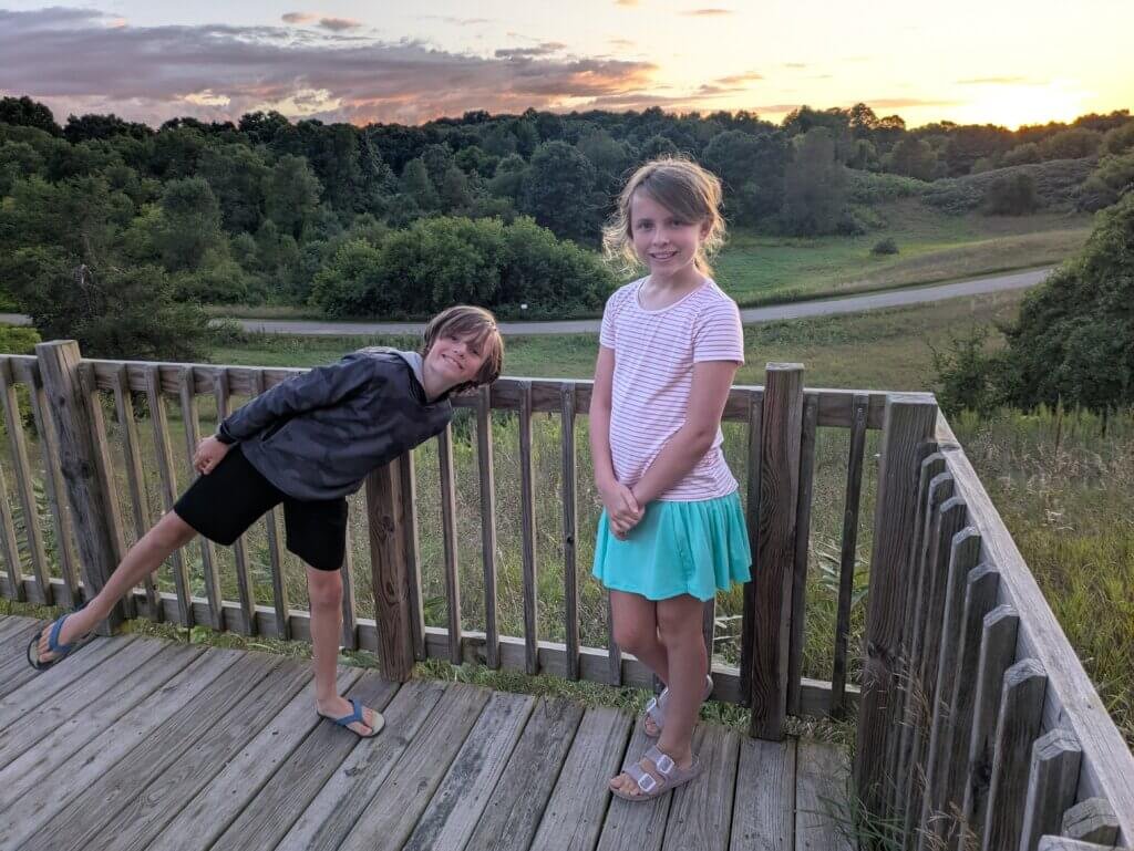 2 kids on deck in front of woods, field and road